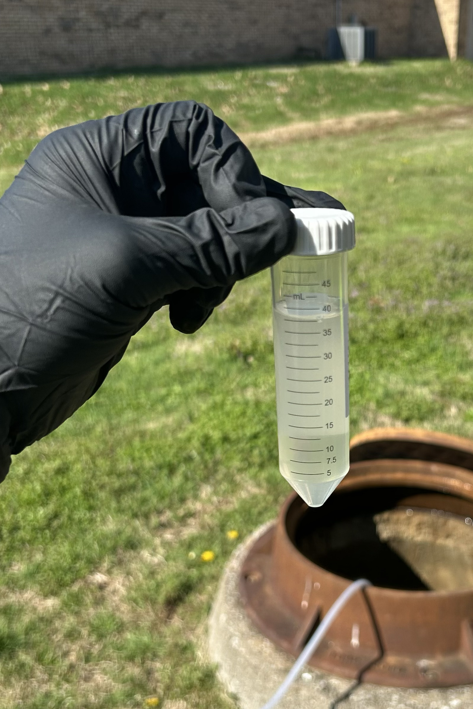 A gloved hand holding a sample tube above an open manhole.